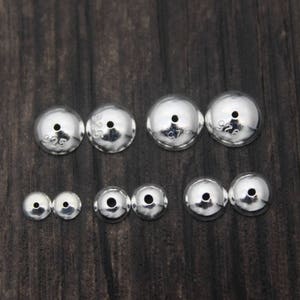 May include: A set of ten silver-toned metal beads with a hole in the center. The beads are different sizes and are arranged in two rows on a dark wood surface. The beads are marked with the number "925" indicating they are made of sterling silver.