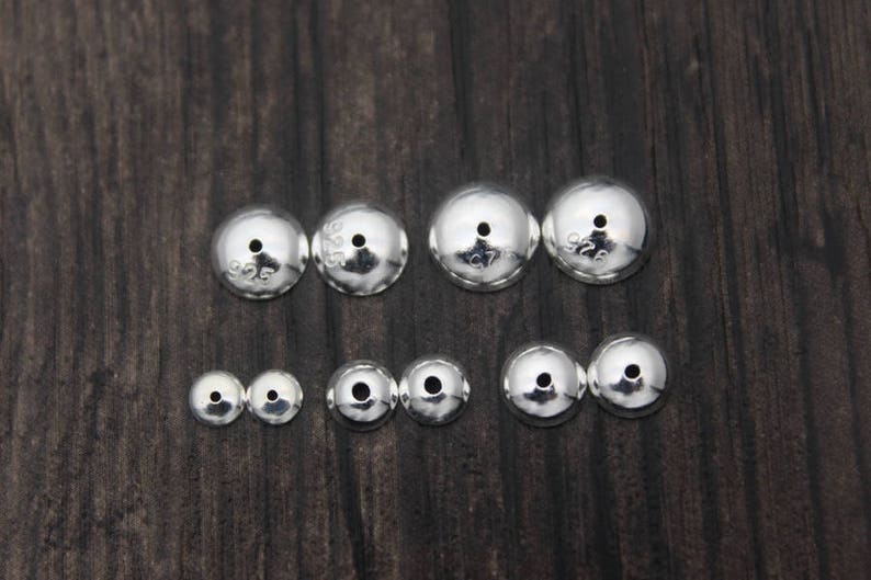 May include: A set of ten silver-toned metal beads with a hole in the center. The beads are different sizes and are arranged in two rows on a dark wood surface. The beads are marked with the number "925" indicating they are made of sterling silver.