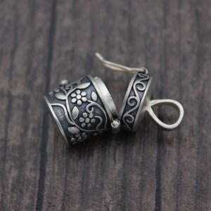 May include: A silver charm with a floral design and a hinged lid. The charm is open and the inside is visible. The charm is on a wooden surface.