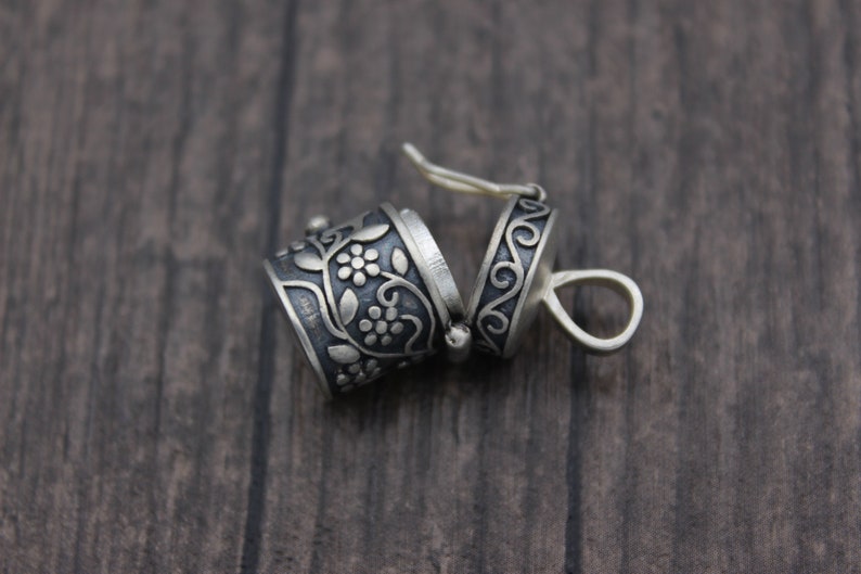 May include: A silver charm with a floral design and a hinged lid. The charm is open and the inside is visible. The charm is on a wooden surface.