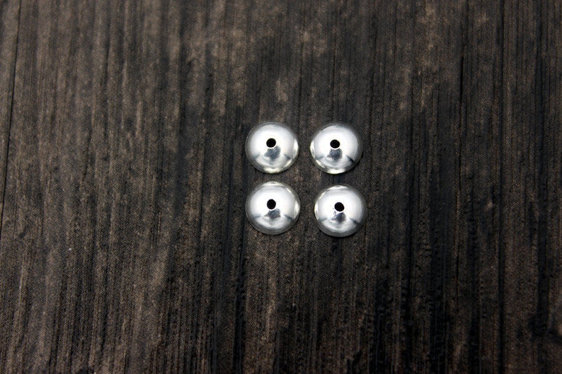 May include: Four silver-colored metal bead caps, arranged in a square pattern on a dark wood surface.