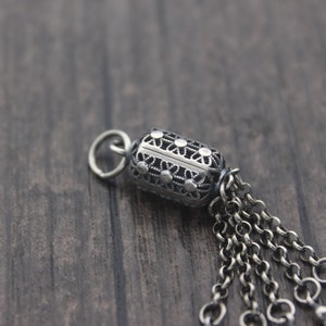May include: A silver charm with an intricate design and a chain tassel. The charm is shaped like a cylinder and has a loop at the top for attaching to a bracelet or necklace.