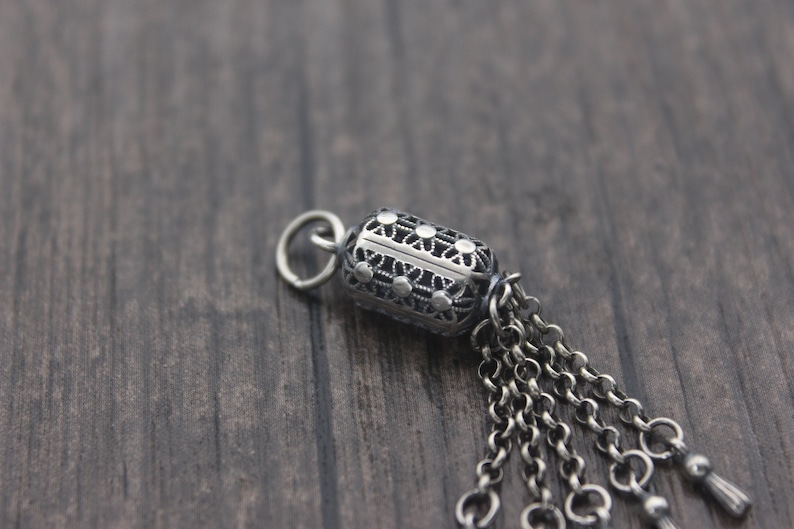 May include: A silver charm with an intricate design and a chain tassel. The charm is shaped like a cylinder and has a loop at the top for attaching to a bracelet or necklace.