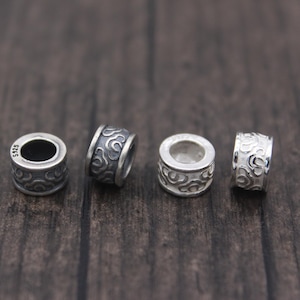 May include: Four small, silver-colored cylindrical beads with an engraved swirling pattern. The beads are arranged in a row on a dark wooden surface. One bead has the marking "S925". These beads are likely for jewelry making.