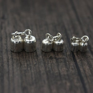 May include: Close-up of several pairs of small, silver-colored dumbbell-shaped charms. Each charm has a loop at the top, connected by a small ring. The charms are arranged on a dark wooden surface.