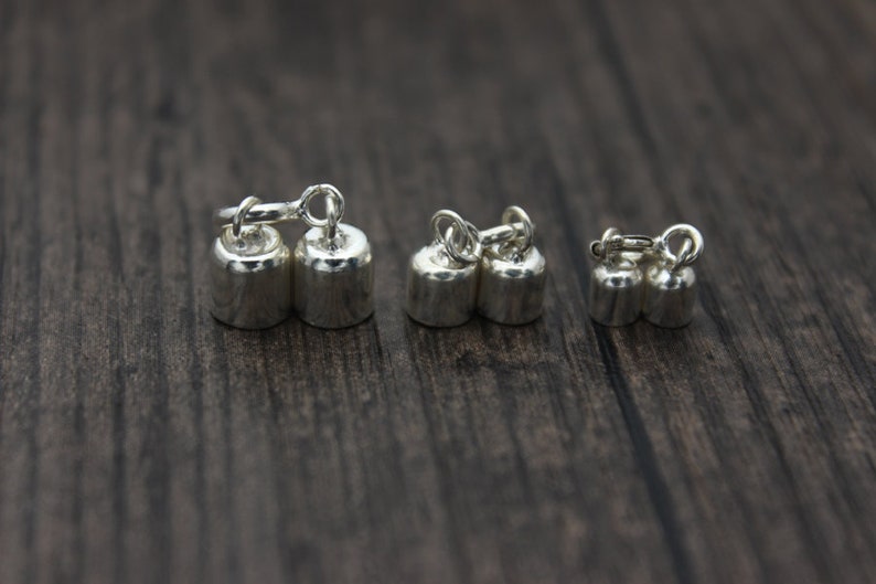 May include: Close-up of several pairs of small, silver-colored dumbbell-shaped charms. Each charm has a loop at the top, connected by a small ring. The charms are arranged on a dark wooden surface.