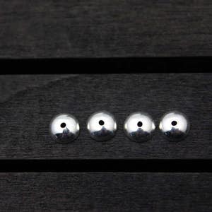 May include: Four silver metal beads with a small hole in the center of each bead. The beads are arranged in a row on a dark wooden surface.