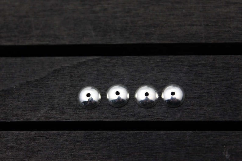 May include: Four silver metal beads with a small hole in the center of each bead. The beads are arranged in a row on a dark wooden surface.