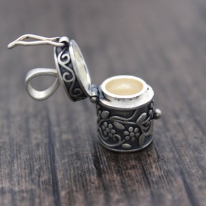 May include: A silver charm in the shape of a small, ornate box with a hinged lid. The box is decorated with a floral design and has a loop for attaching to a necklace or bracelet.