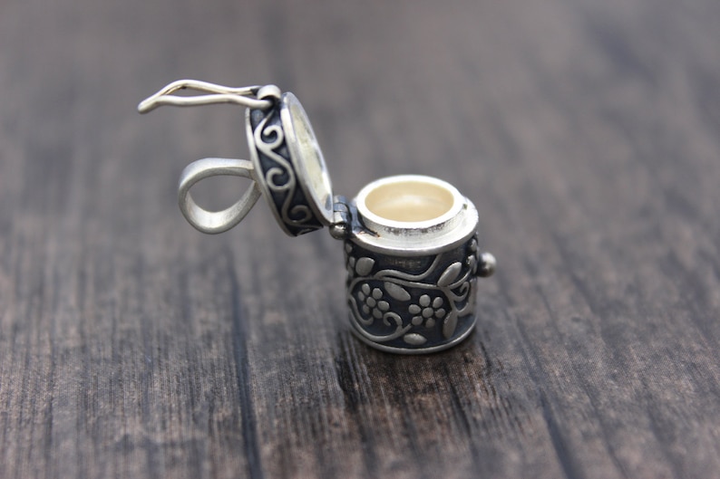 May include: A silver charm in the shape of a small, ornate box with a hinged lid. The box is decorated with a floral design and has a loop for attaching to a necklace or bracelet.