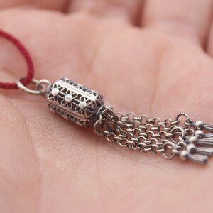 May include: A silver charm with a detailed design and a tassel of chains. The charm is attached to a red cord.