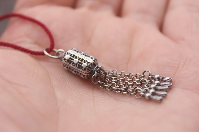 May include: A silver charm with a detailed design and a tassel of chains. The charm is attached to a red cord.