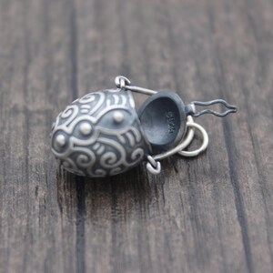 May include: A silver charm in the shape of a small, ornate bucket with a hinged lid. The bucket is decorated with a swirling pattern and has a small loop for attaching to a chain.