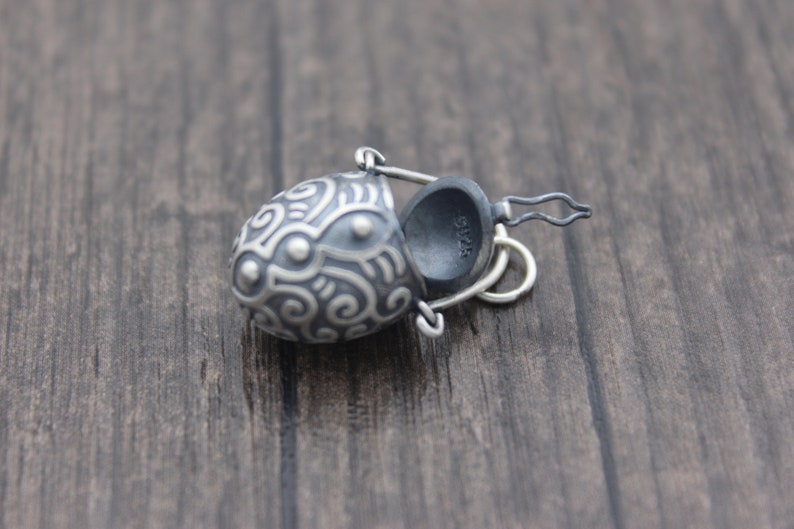 May include: A silver charm in the shape of a small, ornate bucket with a hinged lid. The bucket is decorated with a swirling pattern and has a small loop for attaching to a chain.