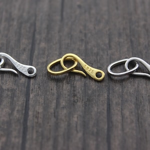 May include: Three silver-toned metal clasps with a hook design. Each clasp is inscribed with "S925" on the side. The clasps are arranged in a row on a wooden surface.
