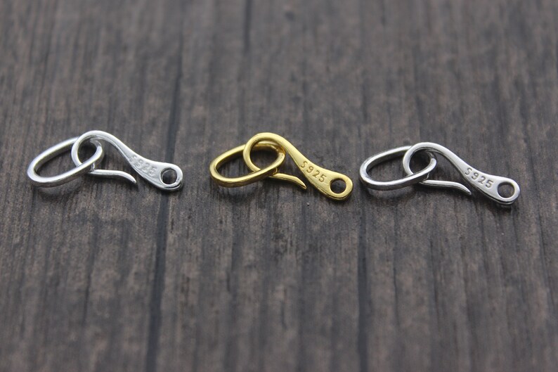 May include: Three silver-toned metal clasps with a hook design. Each clasp is inscribed with "S925" on the side. The clasps are arranged in a row on a wooden surface.