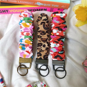 May include: Three fabric keychains with varied patterns: a colourful floral print, a leopard print, and a red and black abstract design. Each keychain has a metal clasp and a ring for keys. The keychains are on a white surface.