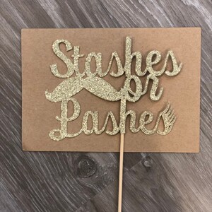 May include: Gold glitter cake topper with the words "Stashes or Lashes" and a glitter mustache.