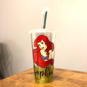 May include: A silver glitter tumbler with a green straw. The tumbler features a cartoon image of Ariel, the Little Mermaid, with the name "Sophia" written below her.