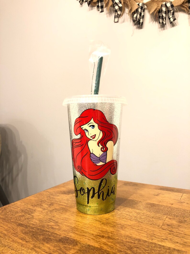 May include: A silver glitter tumbler with a green straw. The tumbler features a cartoon image of Ariel, the Little Mermaid, with the name "Sophia" written below her.