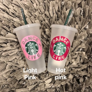 May include: Two clear plastic reusable cups with lids and green straws. The cups have a pink and red design with the words "Dance Life" and a stylized mermaid logo. The cups are on a brown and white fuzzy surface. The text below the cups reads "Light Pink" and "Hot Pink".