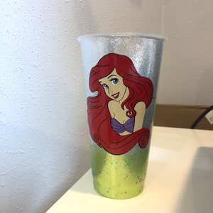 May include: A silver glitter tumbler with a green glitter bottom. The tumbler features a printed image of Ariel, the Little Mermaid, with red hair and a purple seashell bra.