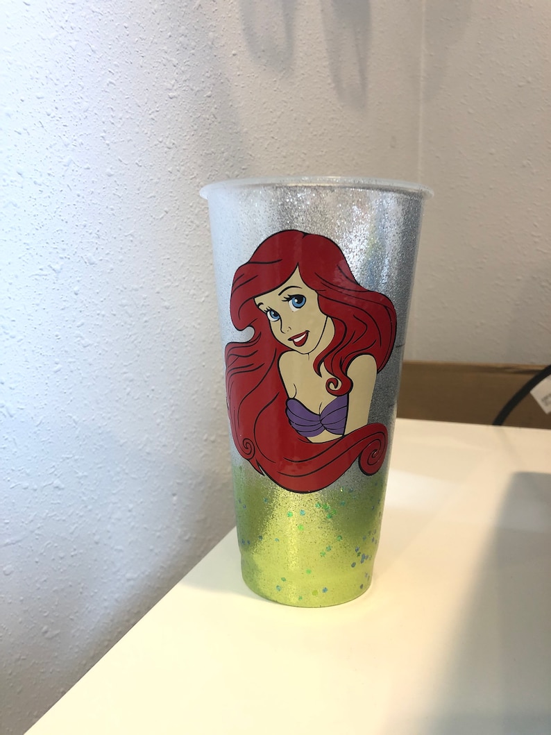May include: A silver glitter tumbler with a green glitter bottom. The tumbler features a printed image of Ariel, the Little Mermaid, with red hair and a purple seashell bra.