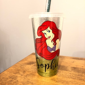May include: A silver glitter tumbler with a green straw. The tumbler has a picture of Ariel from The Little Mermaid and the name "Sophia" written in black glitter.
