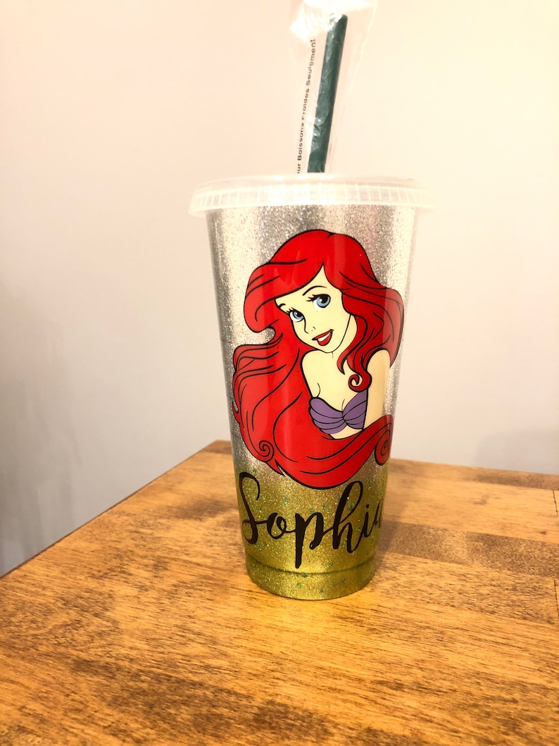 May include: A silver glitter tumbler with a green straw. The tumbler has a picture of Ariel from The Little Mermaid and the name "Sophia" written in black glitter.