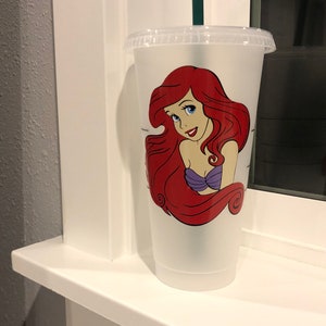 May include: A clear plastic reusable cup with a red-haired mermaid character with a purple top. The mermaid has blue eyes and a smile. The cup has a black lid and a green straw.