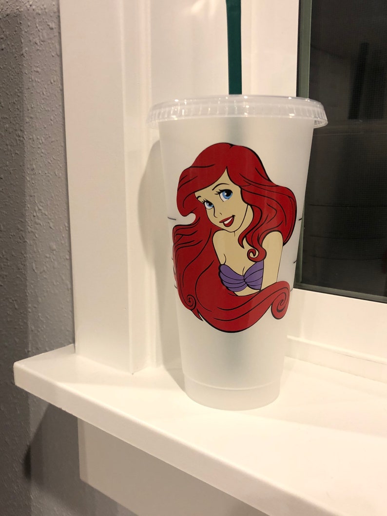 May include: A clear plastic reusable cup with a red-haired mermaid character with a purple top. The mermaid has blue eyes and a smile. The cup has a black lid and a green straw.