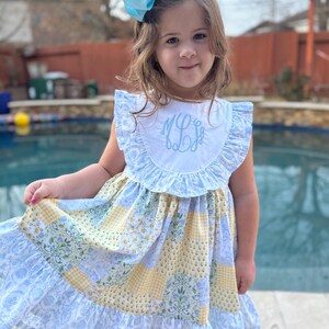 May include: A little girl wearing a blue and yellow patchwork dress with a white collar and blue ruffle trim. The dress has a monogram on the collar. She is wearing a blue bow in her hair.