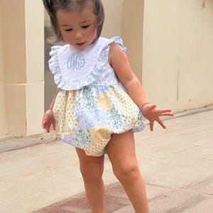 May include: A little girl wearing a blue and yellow floral print romper with a white ruffle collar and a monogrammed "GGS" on the front. She is wearing her hair in pigtails with blue bows.