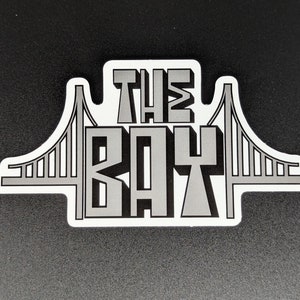 May include: A white sticker with a gray outline of the Golden Gate Bridge and the text "THE BAY" in gray letters.