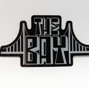 May include: Black and silver sticker with the text "THE BAY" and a stylized image of the Golden Gate Bridge.