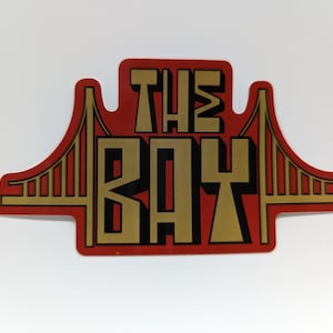 May include: A red sticker with the words "THE BAY" in gold block letters. The design incorporates a stylized image of the Golden Gate Bridge in gold, with a red background. The sticker is rectangular with rounded edges.