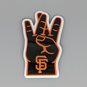 May include: A black and orange foam hand with the San Francisco Giants logo. The hand is in a three-finger gesture, with the fingers and outline in orange. The logo is in the center of the palm.
