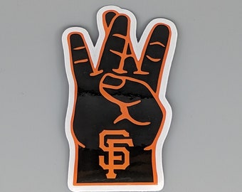 May include: A black and orange foam hand with the San Francisco Giants logo. The hand is in a three-finger gesture, with the fingers and outline in orange. The logo is in the center of the palm.