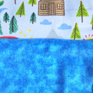 May include: A blue and white fabric with a camping theme. The fabric features a cabin, trees, mountains, and stars. The bottom half of the fabric is a solid blue colour.