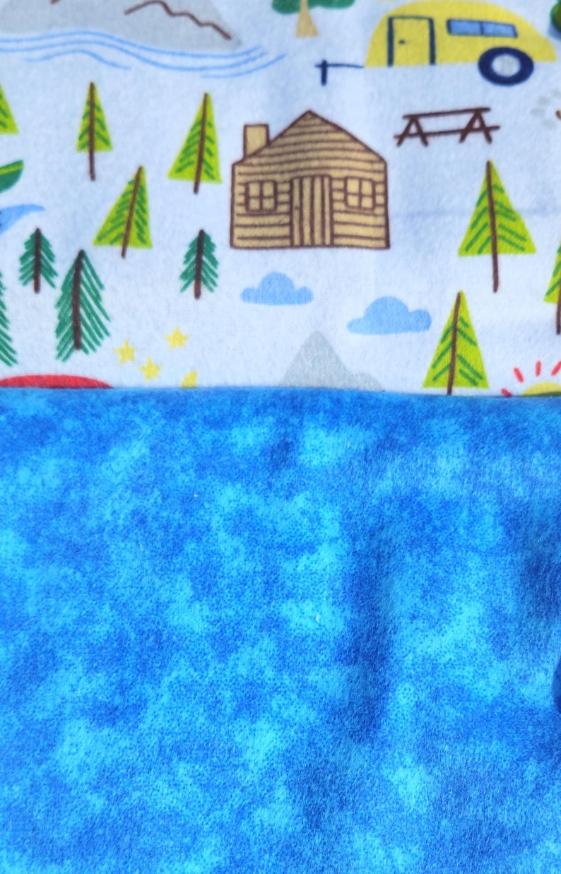 May include: A blue and white fabric with a camping theme. The fabric features a cabin, trees, mountains, and stars. The bottom half of the fabric is a solid blue colour.