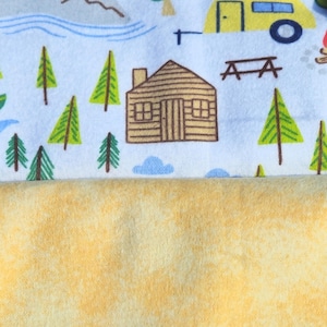 May include: A white fabric with a repeating pattern of a cabin, trees, mountains, and a yellow camper. The fabric is folded over a yellow fleece fabric.