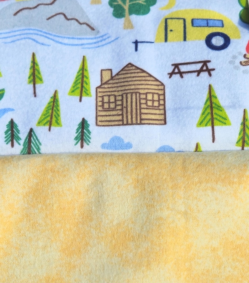 May include: A white fabric with a repeating pattern of a cabin, trees, mountains, and a yellow camper. The fabric is folded over a yellow fleece fabric.