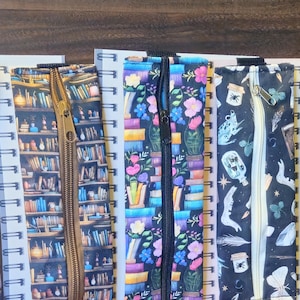 May include: A collection of colorful journal pen holders with various patterns, including books, floral designs, and celestial motifs. Each holder has a zipper closure and is attached to a spiral notebook. The text "JOURNAL PEN HOLDER" is visible.