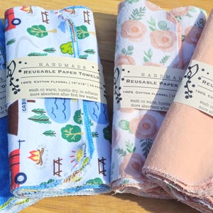 May include: Four handmade reusable paper towels, each with a different pattern. The towels are made of 100% cotton flannel and measure 10x12 inches. The towels are folded and stacked on a wooden surface. The towels are labelled "Handmade Reusable Paper Towels 100% Cotton Flannel 10x12" and have a blue, pink, floral, and camping themed design.
