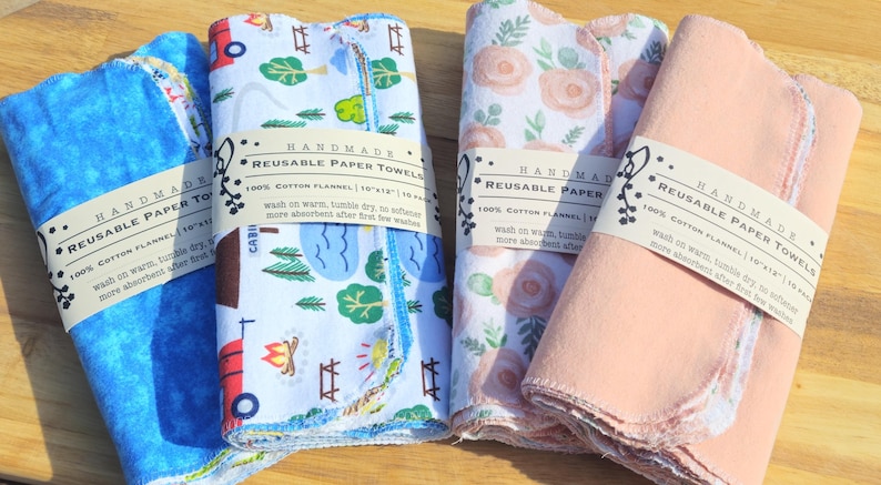 May include: Four handmade reusable paper towels, each with a different pattern. The towels are made of 100% cotton flannel and measure 10x12 inches. The towels are folded and stacked on a wooden surface. The towels are labelled "Handmade Reusable Paper Towels 100% Cotton Flannel 10x12" and have a blue, pink, floral, and camping themed design.