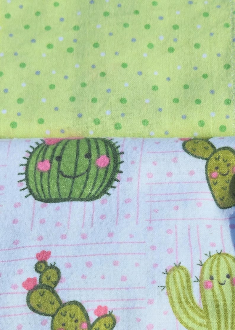May include: A white fabric with a pink and green cactus pattern. The cactus has a smiling face and pink cheeks. The fabric has a light green polka dot pattern on the top.