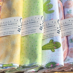 May include: Ten stacks of handmade reusable paper towels with various colourful patterns, including cacti, flowers, and geometric designs. Each stack has a label that reads "Handmade Reusable Paper Towels 10" x 10" / 10 in pack.  Wash, wring, dry, and use again and again.  No softener.
