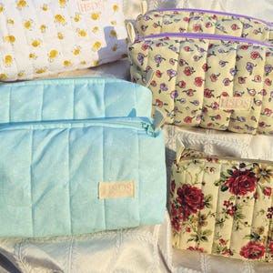 May include: Several quilted cosmetic bags in various patterns. One is light blue, another has a bee pattern, and others feature floral designs. Each bag has a zipper closure and a small label with the letters "HSDS".