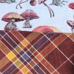 May include: A white fabric with a mushroom print pattern. The fabric is folded over a brown and orange plaid fabric.