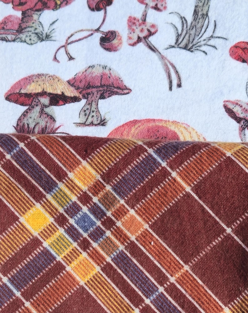 May include: A white fabric with a mushroom print pattern. The fabric is folded over a brown and orange plaid fabric.